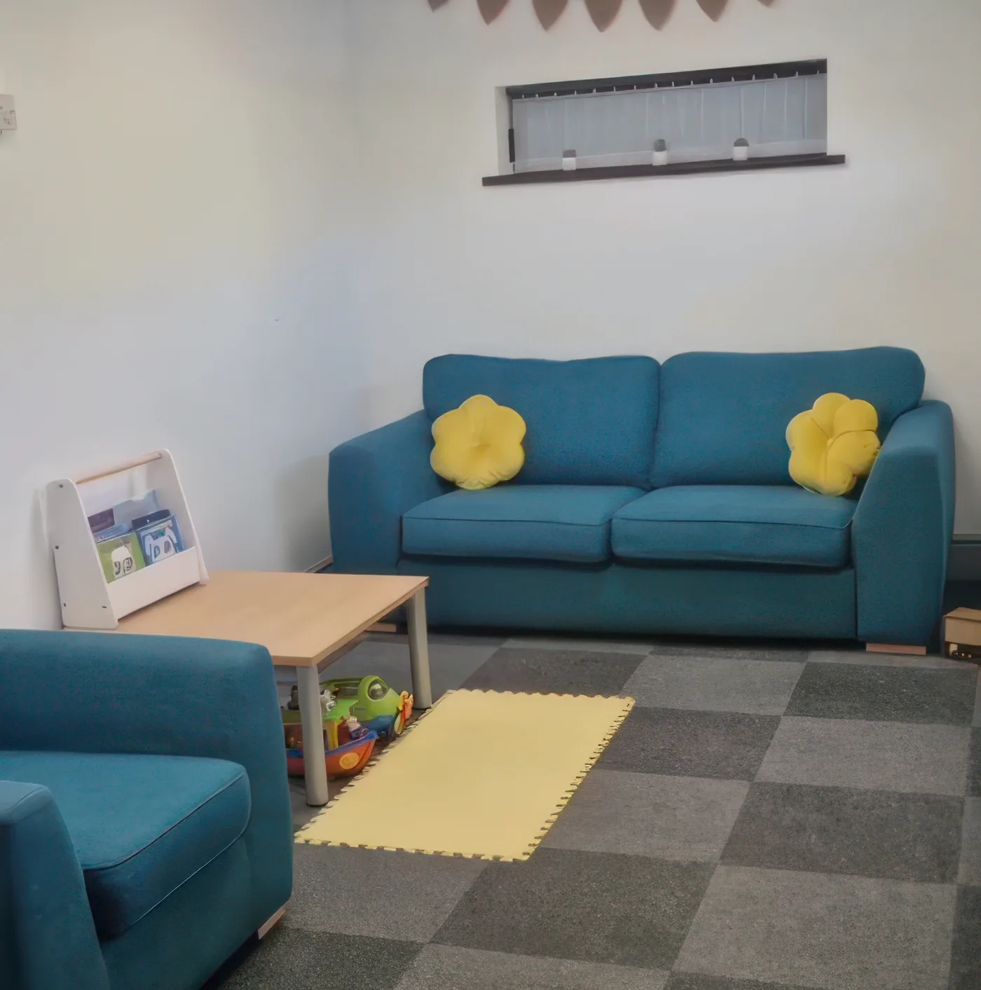 The waiting area at the Ossett clinic: a teal sofa and armchair with a low table, a rack of children's books, a box of toys and a soft play mat on carpet tiles